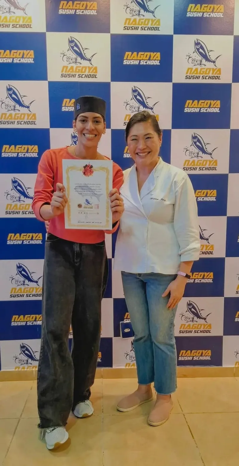 Joy ao lado de uma mulher de camisa branca, com um certificado nas mãos, diante do painel da Nagoya Sushi School