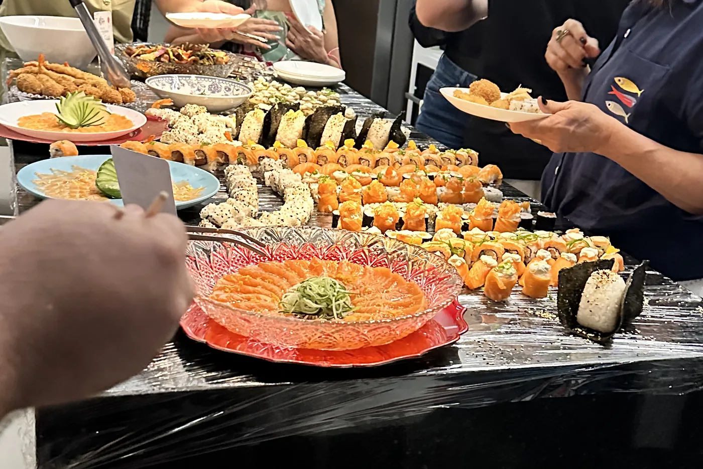 Mesa de evento com sashimi de salmão, uramakis e hot rolls, enquanto convidados se servem